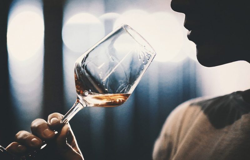 Closeup of unrecognizable adult man slightly swirling a glass of rose wine under his nose and trying to catch bouquet. She's in a wine cellar, there are blurry metal tanks in background. Backlit