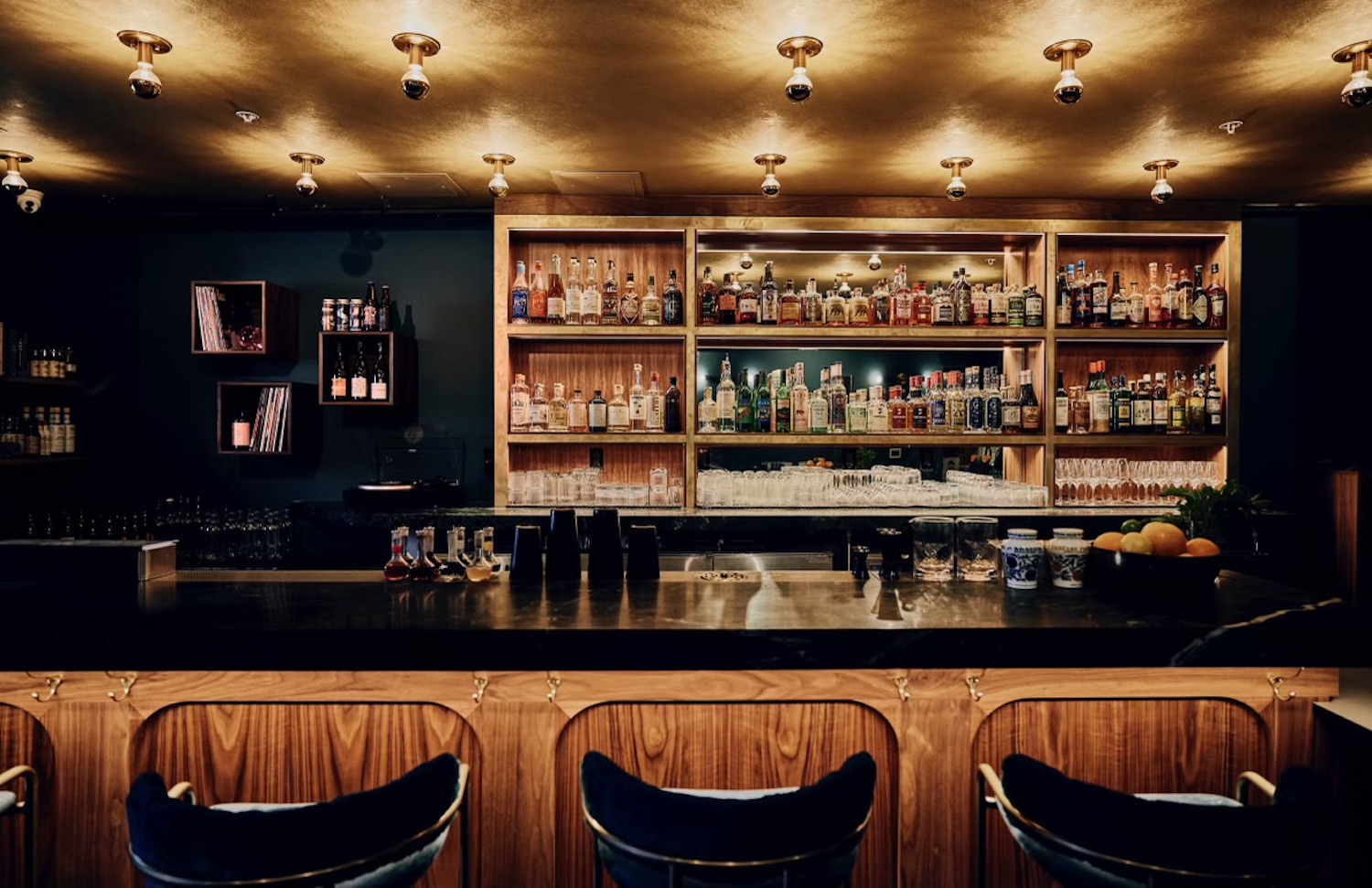 Bar area with light wood fixtures, barstools and shelves full of bottles behind the wall