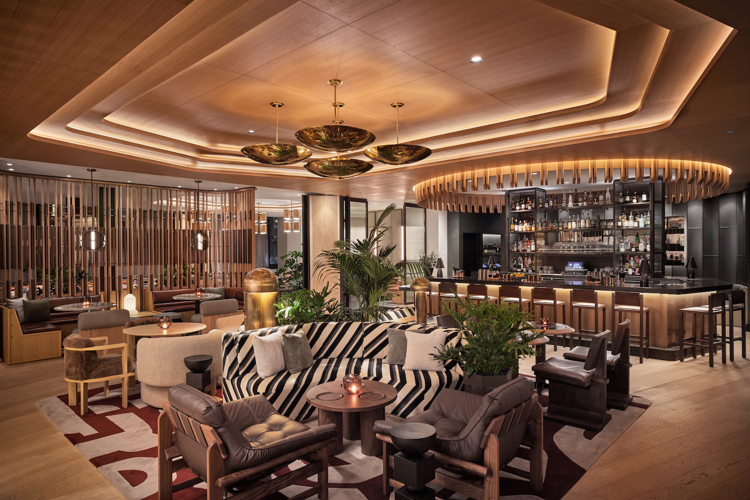 Hotel bar area with several large printed couches under a hanging chandelier and in front of large dark-colored bar with a tall bar rack in the center behind it