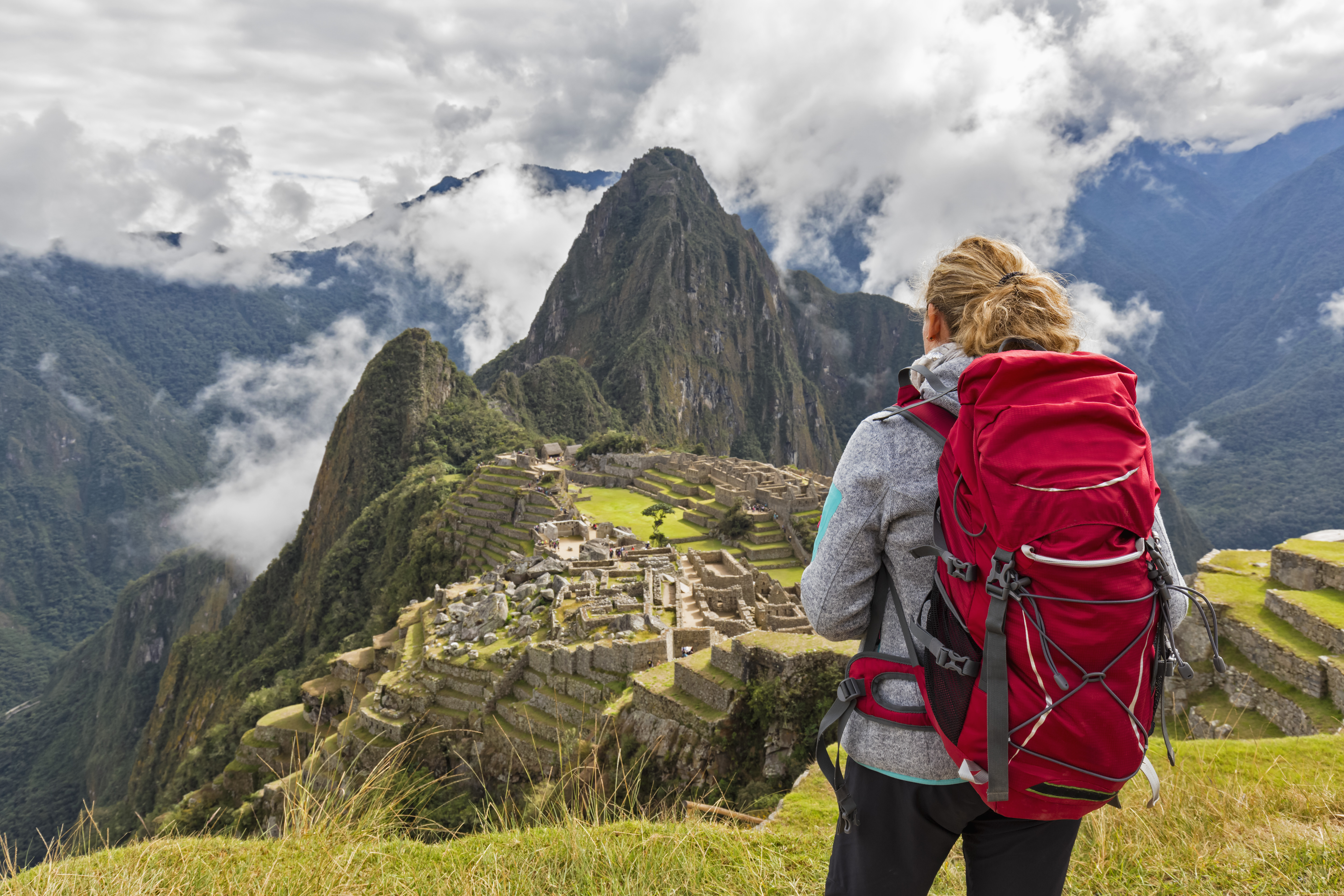 Südamerika, Peru, Anden, Urubamba, Urubambatal, Region Cusco, Aguas Calientes, Touristin (w) mit rotem Rucksack blickt auf die Inka Ruinenstadt Machu Picchu mit dem Berggipfel Huayna Picchu