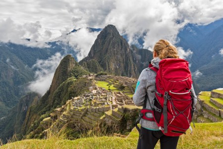 Südamerika, Peru, Anden, Urubamba, Urubambatal, Region Cusco, Aguas Calientes, Touristin (w) mit rotem Rucksack blickt auf die Inka Ruinenstadt Machu Picchu mit dem Berggipfel Huayna Picchu