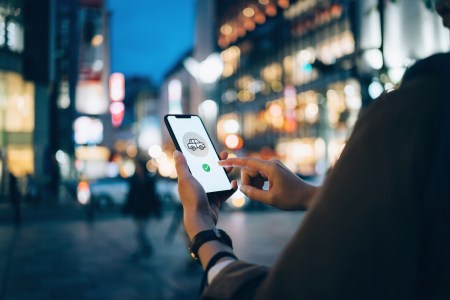 Young woman using mobile app on smart phone to arrange taxi ride in downtown city street, with illuminated city traffic scene as background
