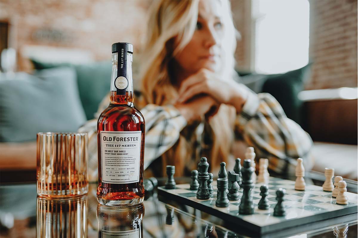 Old Forester Master Taster Jackie Zykan sitting at a table with a chess set and a bottle and glass of Old Forester 117 High Angels' Share
