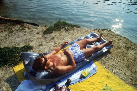 A sunburned man lies shirtless on a pool float by a lake, tanning under direct sunlight.