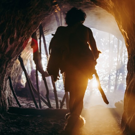 Primeval Caveman in Animal Skin and Fur Holds Stone Tipped Spear Comes out of His Cave into Prehistoric Forest Ready to Hunt. Neanderthal Going Hunting into the Jungle. Shot with Warm Filter.