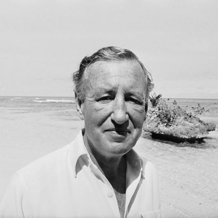 A black and white photo of author Ian Fleming on a beach. We take a look at how his wartime espionage work influenced writing his James Bond books.