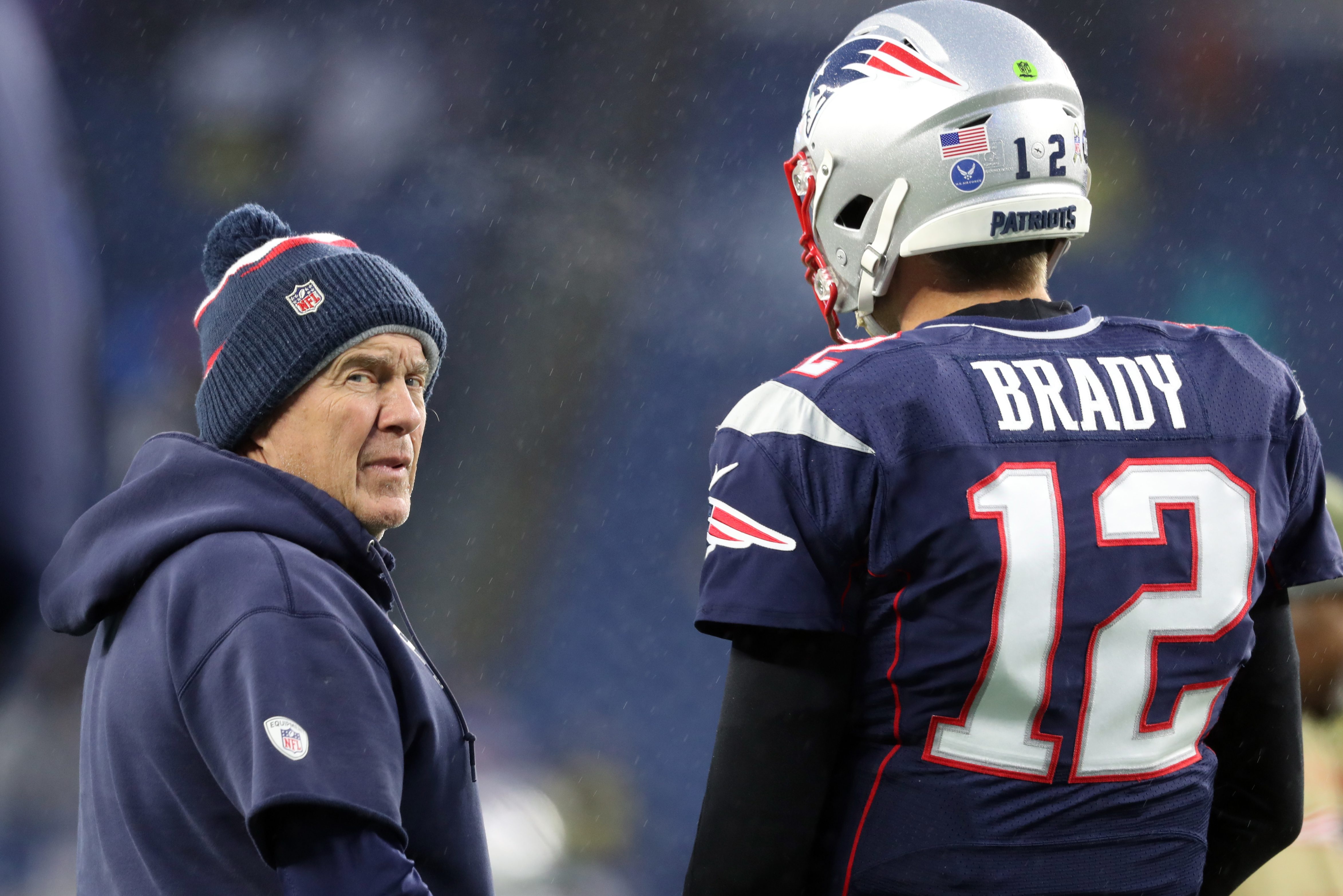 New England Patriots head coach Bill Belichick in a beanie and Tom Brady in full uniform with a helmet talk on the NFL field in 2019