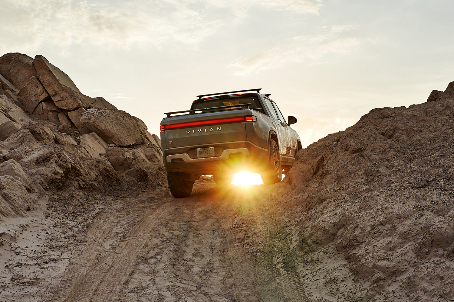 The Rivian R1T electric pickup truck shown from the rear driving over a rocky dirt hill at sunset