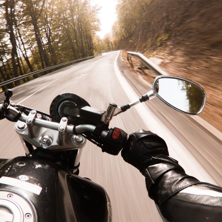 The handlebars and side mirror of a motorcycle as it speeds down a winding road in the woods. A motorcycle just a record for the Cannonball Run, an illegal nationwide road race.