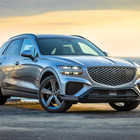 The Genesis GV70 SUV with the headlights on sitting still with a beach and sunset in the background