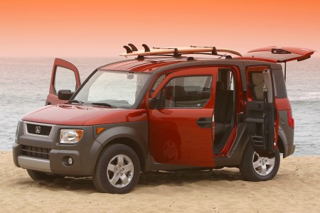 An orange and brown Honda Element SUV with all the doors open and a surfboard on top sitting on the beach at sunset