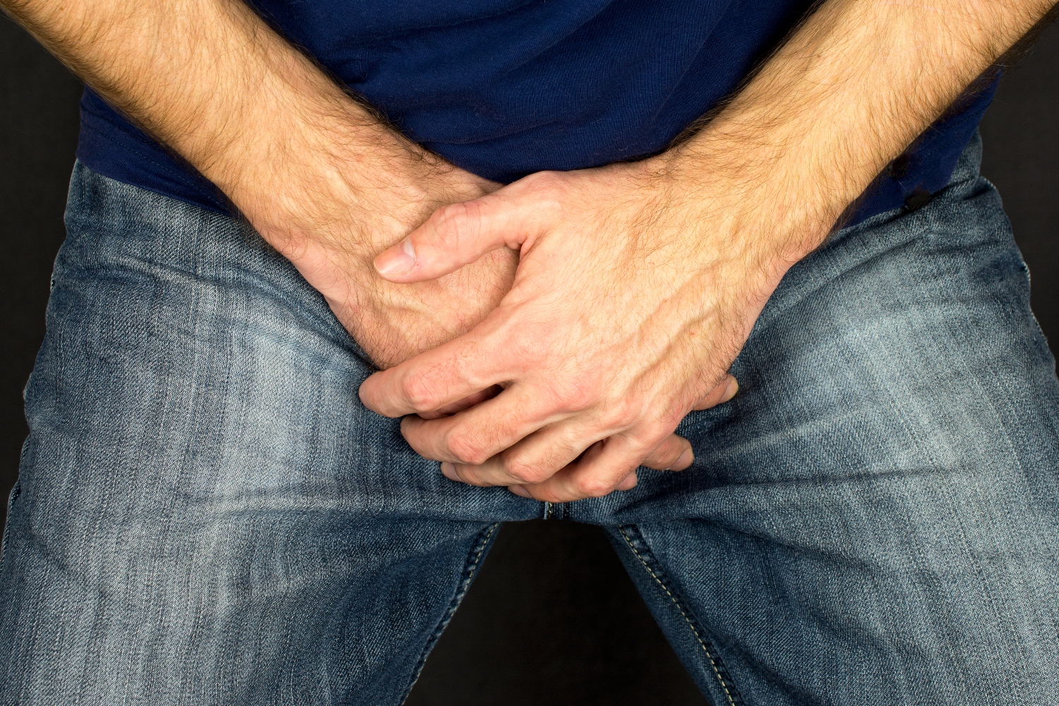 Close up photo shows a man holding his hands folded over his groin while wearing blue jeans