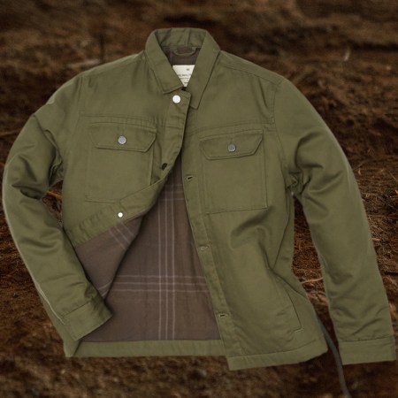 Tentree's Treeline Canvas Unisex Jacket in olive green on a dirt background. The outerwear is supposedly 99% compostable.