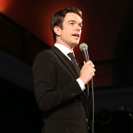 Comedian John Mulaney wearing a suit and tie and holding a microphone at the American Museum of Natural History's 2019 gala