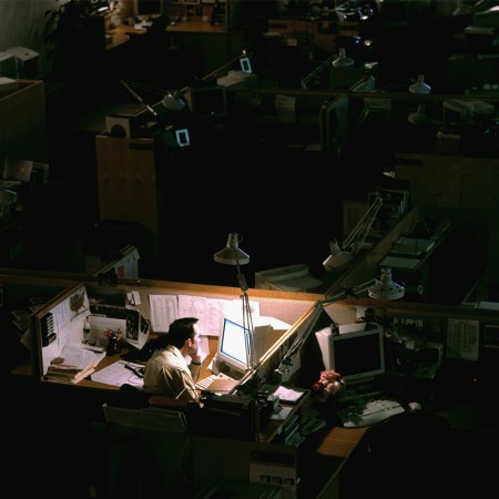 A man sitting at a lit cubicle in a dark office.