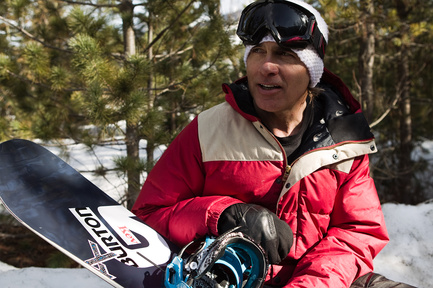 Jake Burton with a beanie and goggles on sitting in the snow holding a snowboard