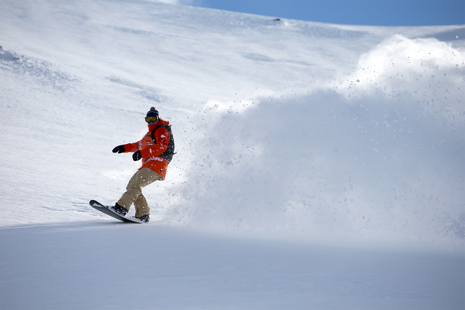 Jake Burton in an orange jacket snowboarding down a mountain with snow flying behind him