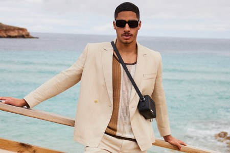 a model in a white suit, shirt and black leather bag standing in front of the ocean, courtesy of Mytheresa