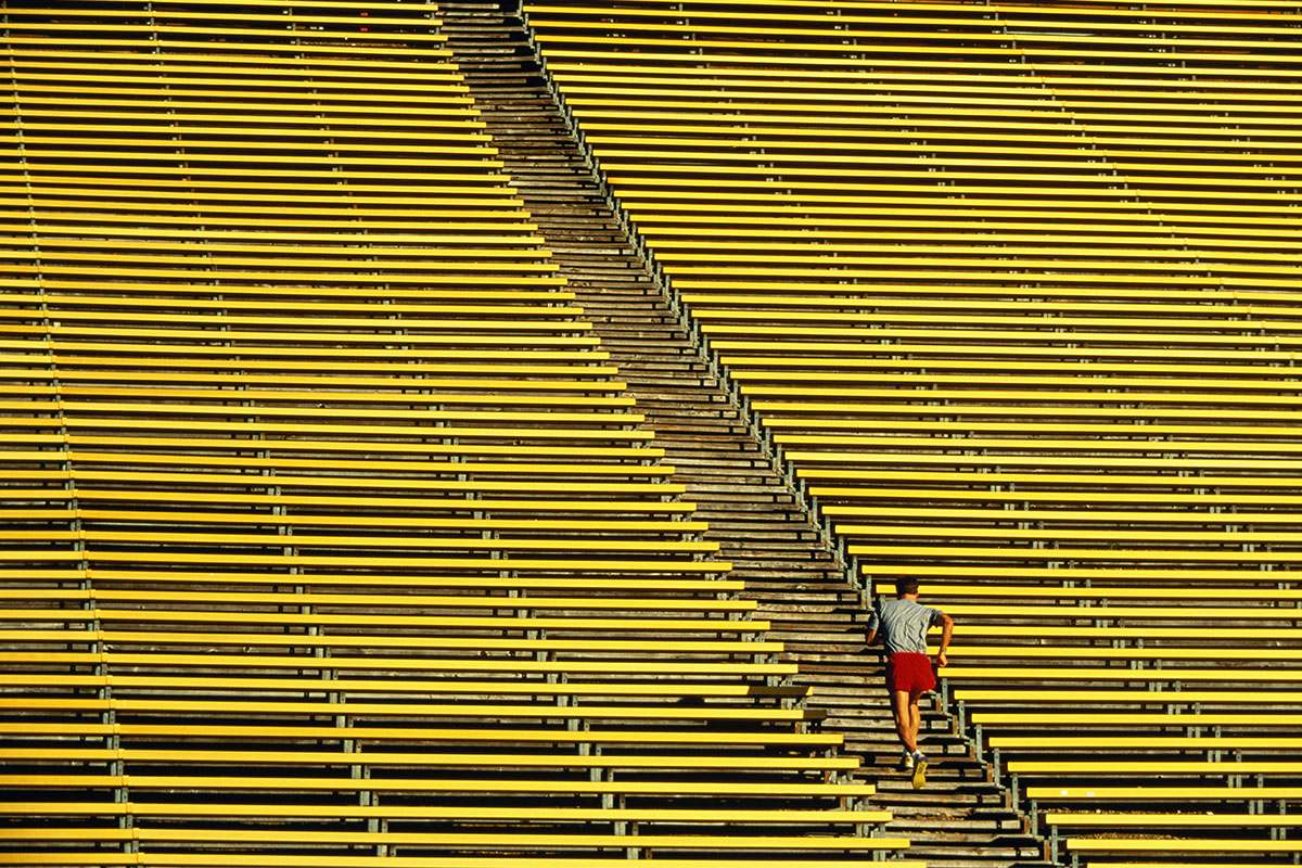 A man in red shorts and a grey shirt running up yellow steps in a football stadium.