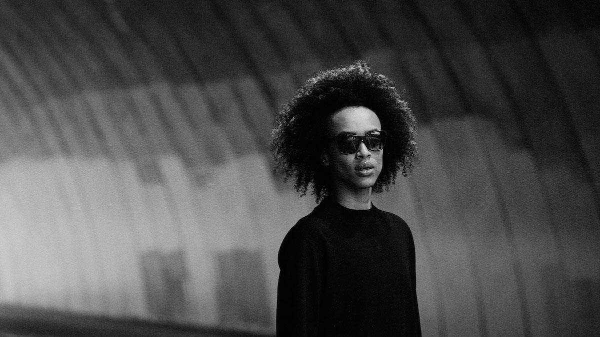 a black and white photo of a model in a black shirt standing in a tunnel wearing a pair of the District Vision x Reigning Champ eyewear collection