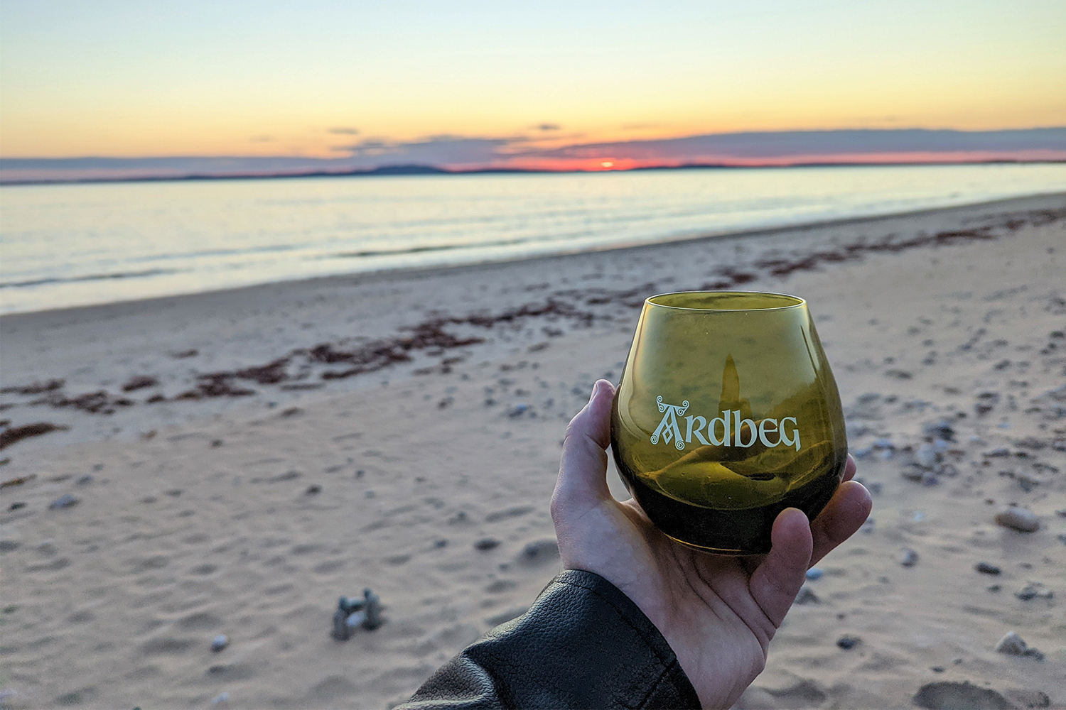 A hand holding a whisky tasting glass on the beach of Islay, Scotland, during the island's 2022 Feis Ile