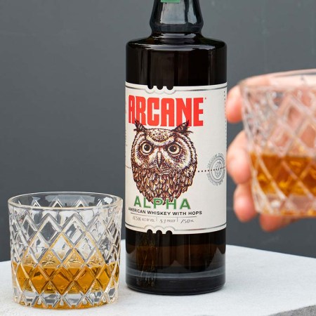 A bottle of Arcane Alpha and two glasses of whiskey on a table