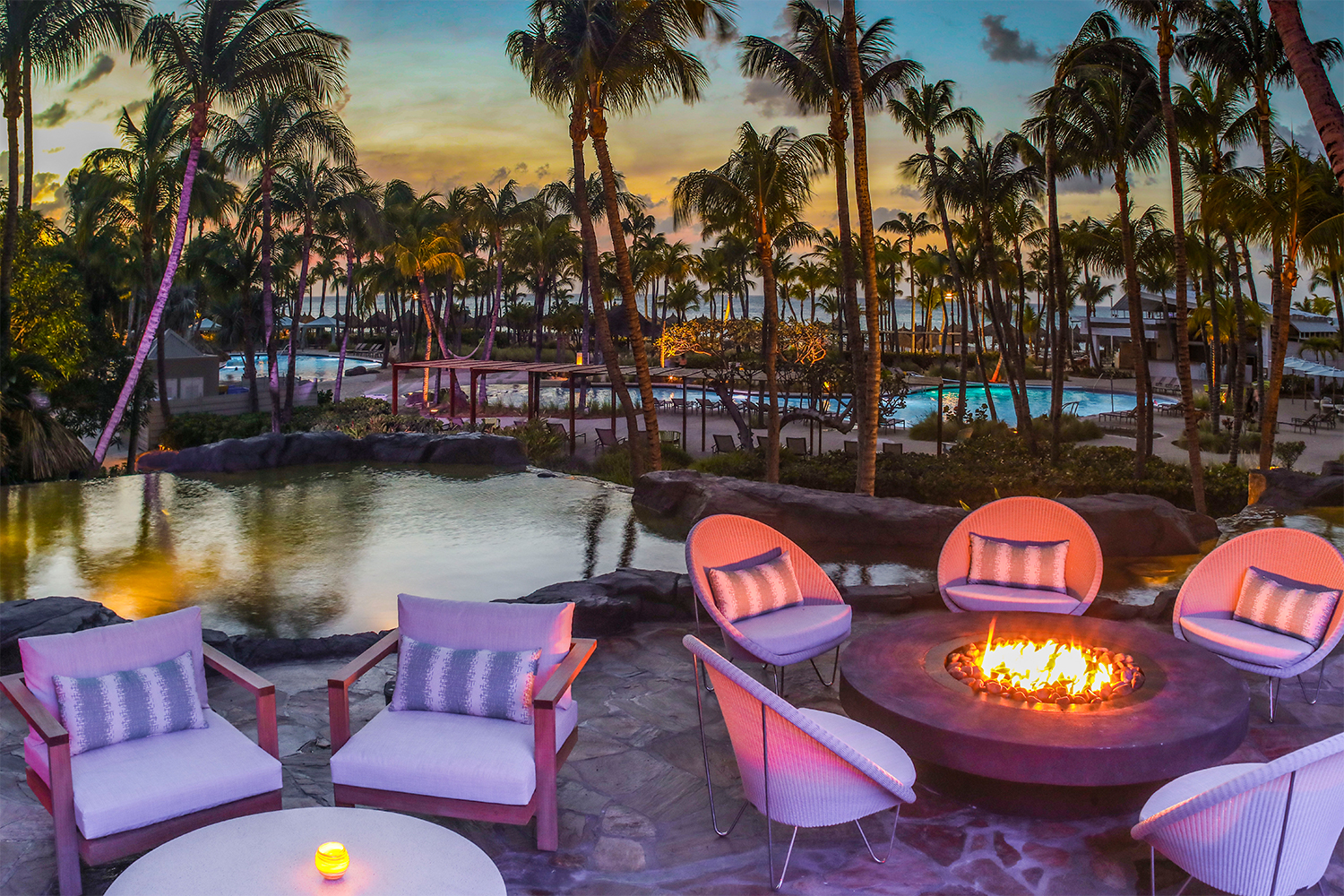 A sunset over the ocean in the background while chairs sit around a firepit at the Hilton Aruba hotel