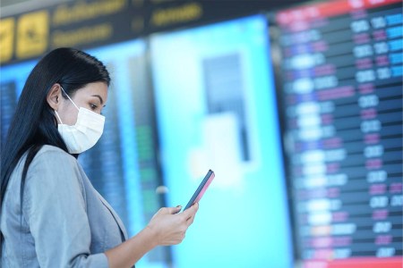 Woman wearing protective face mask checking flight schedule on mobile phone while standing in front of the arrival departure board. A new Twitter scam has fraudulent airline accounts attempting to take money from passengers