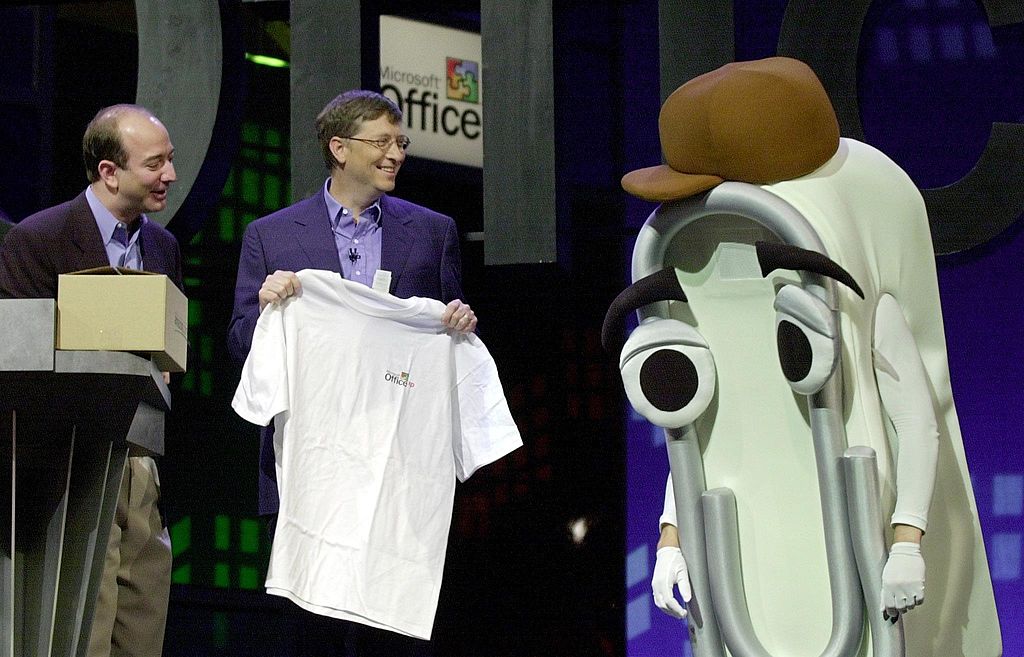 Bill Gates presents a T-shirt as a retirement gift to "Clippy," a person dressed as the Microsoft Office Assistant, as Jeff Bezos looks on