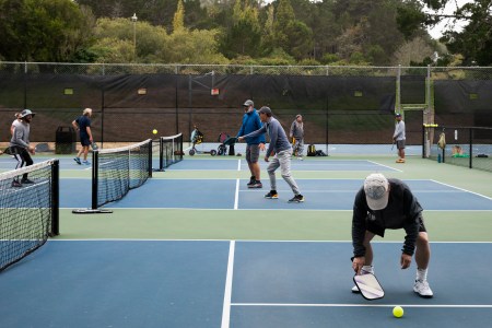 A man bends down on a pickleball court to grab a ball with other players in the background