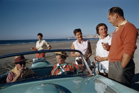 A group of wealthy people gathered around a convertible on a beach.