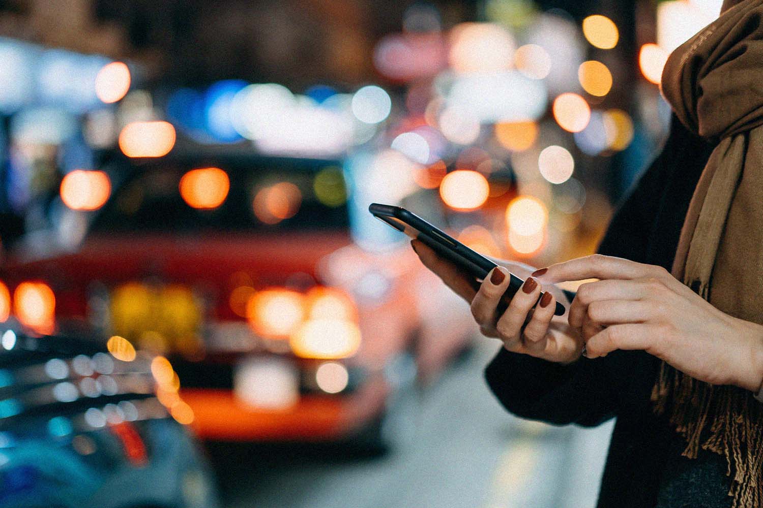 Close up of young woman using mobile app device on smartphone in downtown city street, with illuminated busy city traffic scene during rush hour with traffic congestion in the evening