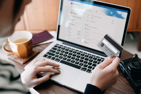 Over the shoulder view of young woman shopping online for flight tickets on airline website with laptop, entering credit card details to make mobile payment at home