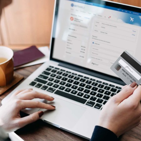 Over the shoulder view of young woman shopping online for flight tickets on airline website with laptop, entering credit card details to make mobile payment at home