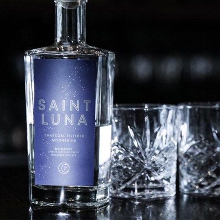 A bottle of Saint Luna moonshine and two glasses on a table.