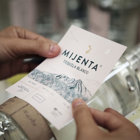 A hand placing the label on a bottle of Mijenta Tequila
