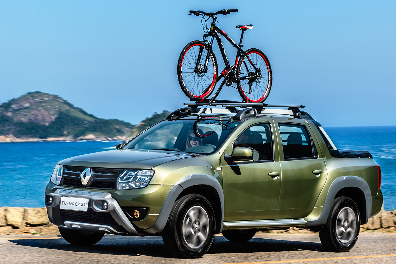Two people inside a green Renault Duster Oroch, a compact pickup truck, with a bike strapped to the top