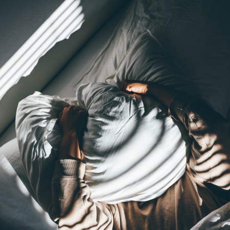 A man holds a pillow over his face as light trickles into his bedroom.