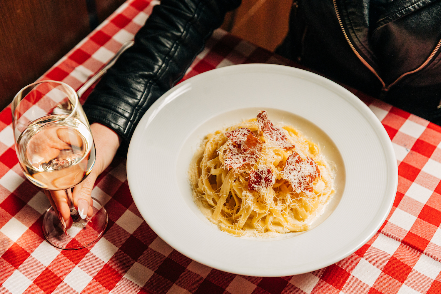 Fettuccine alfredo in a bowl next to a glass of wine on a red and white striped tablecloth