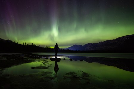 two people standing with their snowmobiles other the northern lights