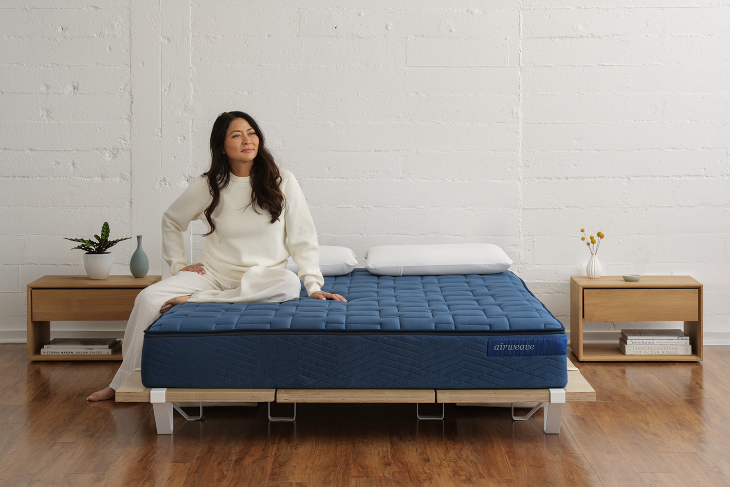 photo of a woman in all white sitting on a blue mattress, which is on a wooden platform, in a simple room with two dressers, wood floors and a blank off white wall