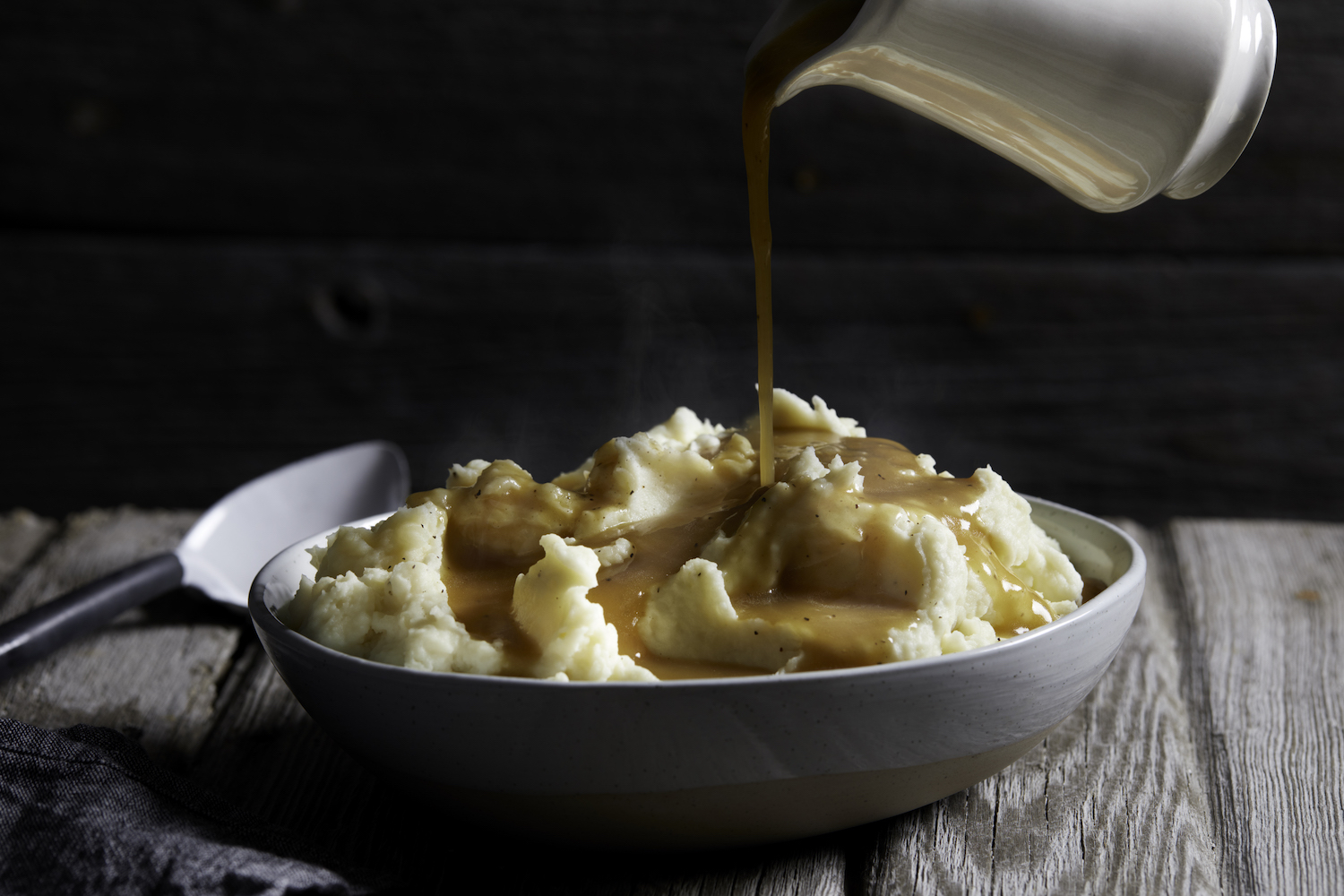 Jug of gravy being poured onto bowl of steaming mashed potatoes