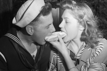 woman and her sailor boyfriend eat from both ends of a hot dog