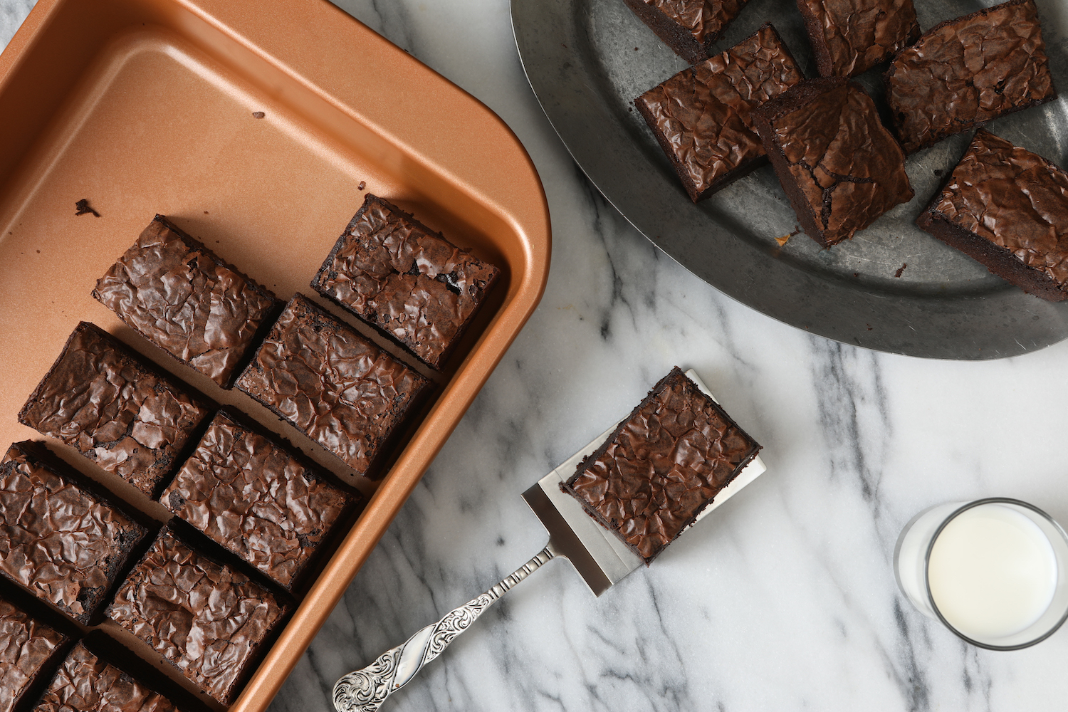 overhead photograph of brownies in a golden baking pan, brownies on an old pewter plate, on a server and a glass of milk.