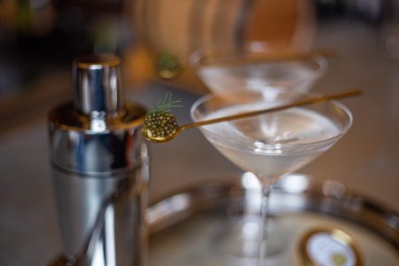 A picture of a martini glass with a spoon of caviar resting on top with a cocktail shaker next to it.