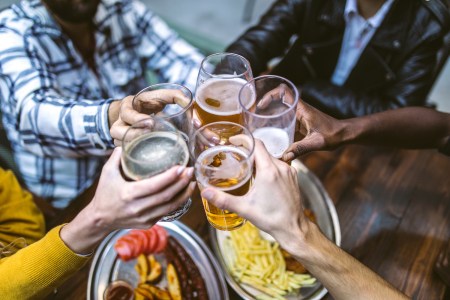 Fried potatoes with sausages, tomato on metal plate at table, unrecognizable people toasting with beer