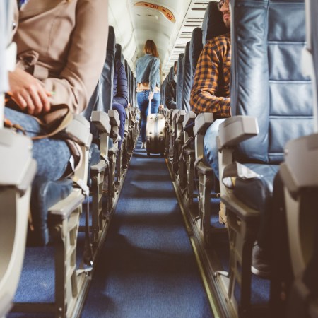 Interior of airplane with people sitting in seats. Passengers with suitcase in aisle looking for seat during flight.