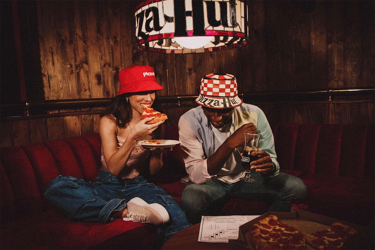 Two models in Pizza Hut bucket hats eating pizza under a branded lamp