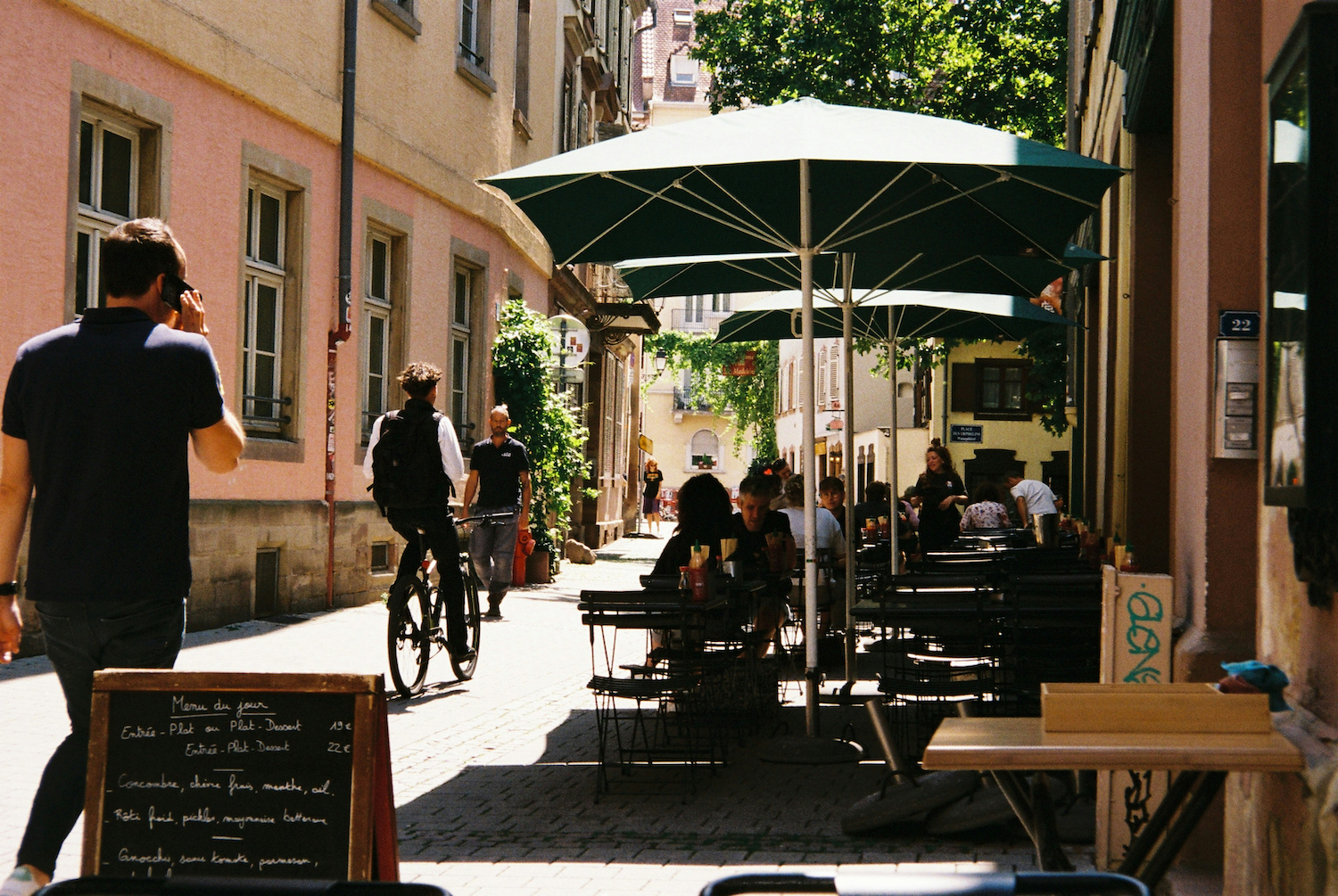 A street in Europe with a man on a bike and patrons at a cafe.