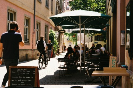 A street in Europe with a man on a bike and patrons at a cafe.